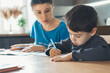 © Strelciuc - Caucasian mother sitting at a table at home helping her son with his homework as he writes notes in a notebook. Mother teaching boy writing. Parent helping