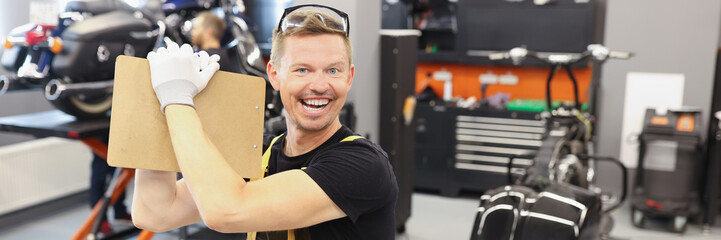 Happy male repairman holding clipboard with documents near motorcycle in car repair shop