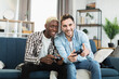 © sofiko14 - Multi ethnic same sex couple sitting together on couch and using wireless joysticks for playing video games. Happy young man spending free time time with fun at home.