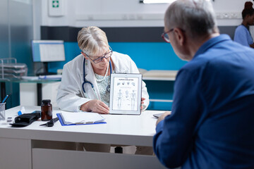 Wall Mural - Physician holding tablet with image of human skeleton to explain diagnosis to old man in cabinet. Doctor and patient looking at bones and spinal cord illustration on device for examination.