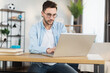 © sofiko14 - Attractive young man in eyewear and casual clothes sitting at domestic workplace and using modern laptop for work. Concept of people, technology and freelance.