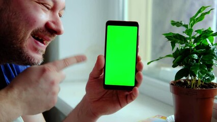A middle-aged man is holding and showing a mobile phone with a green screen in a living room.