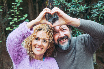 Happy adult couple people in love do hearth symbol gesture with hands together smiling and enjoying relationship. Man and woman leisure with green foliage woods forest in background.