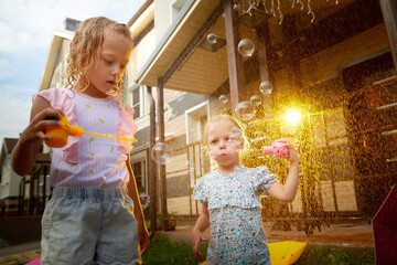 two cute little blonde girls sisters with soap bubbles under rain drops near house and village in a 