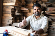 Smiling carpenter showing thumbs up by looking at camera while working at shop - concept of approve, promotion, artisan and wood working.