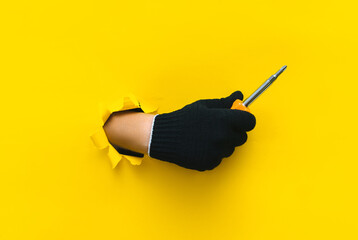 Wall Mural - A right hand in a black cloth knitted glove holds a large orange screwdriver close up. Torn hole in yellow paper. The concept of a worker, labor migrant, a master of his craft. Copy space.