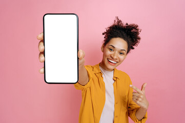 Smartphone mock-up. Excited happy african american girl, holding cellphone in hand with blank white screen for presentation, pointing finger at it, stand over isolated pink background,smiling joyfully