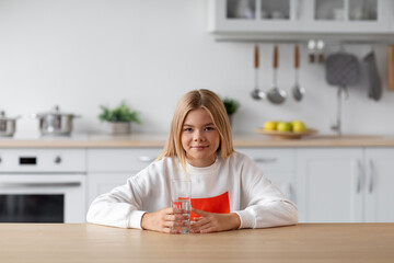 Happy european pretty adolescent blonde girl in casual sits at table with glass of water in minimalist kitchen