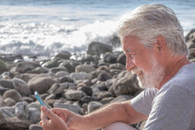 Relaxed And Smiling Adult Bearded Senior Man Sitting On The Rock Beach Using Mobile Phone. White-haired Elderly Grandfather Enjoying Technology And Social And Free Time During Retirement