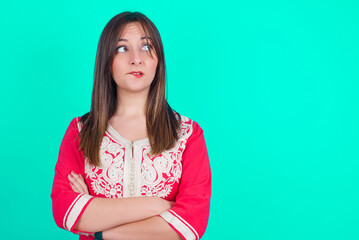 Amazed young beautiful moroccan woman wearing traditional caftan dress over green background bitting lip and looking tricky to empty space.