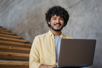 Handsome Indian businessman using laptop computer working in modern  office. Asian student studying, learning language, online education concept. Smiling freelancer copywriter typing looking at camera