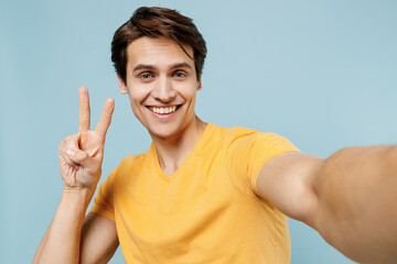 Close up young smiling happy satisfied fun cool man 20s wearing yellow t-shirt doing selfie shot pov on mobile phone show v-sign gesture isolated on plain pastel light blue background studio portrait