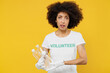 © ViDi Studio - Young confused woman of African American ethnicity wears white volunteer t-shirt gloves hold plastic pet bottle isolated on plain yellow background. Voluntary free work assistance help grace concept.