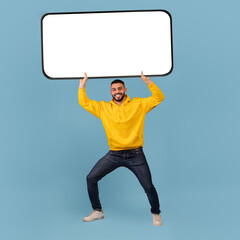 Wall Mural - Millennial arab guy holding big heavy smartphone with white empty screen over blue studio background, mockup