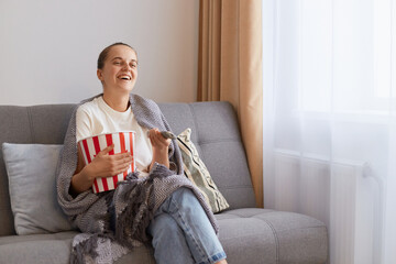 Wall Mural - Indoor shot of woman sitting on sofa wrapped in plaid and watching funny movie and eating popcorn, spending her weekend alone, enjoying comedy film or tv show.