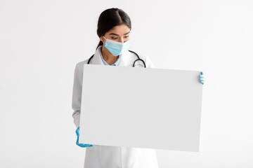 Wall Mural - Young indian woman doctor in uniform, gloves and protective mask shows banner with blank space