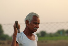 Photo Of An Indian Senior Male Farmer Man Stands In The Field At Sunset With A Shovel On His Shoulder With Wearing A White Vest