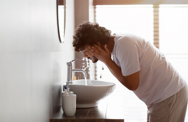 Wall Mural - Young indian man washing face with clear water from tap bending over sink, doing skincare facial routine in bathroom