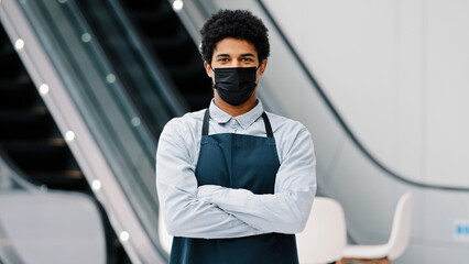 Wall Mural - African american man guy restaurant cafe male worker in medical mask looking camera posing crossing arms waiter salesman wearing apron standing at work space covid pandemic quarantine. Small business