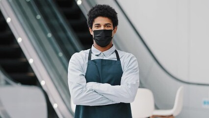 Wall Mural - African american man guy restaurant cafe male worker in medical mask looking camera posing crossing arms waiter salesman wearing apron standing at work space covid pandemic quarantine. Small business