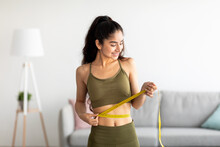 Successful Weight Loss Concept. Fit Young Indian Woman In Sports Clothes Measuring Waist With Tape At Home