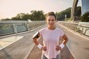 Confident Hispanic sportswoman, female athlete, runner in pink t-shirt, wearing smartphone holder and earphones, smiles looking at camera standing on the city bridge traedmill, relaxing after jog