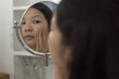 Over the shoulder, close-up shot of a cheerful middle-aged Asian woman looking at the bathroom mirror, checking facial skin condition. Cosmetic and skincare concept.
