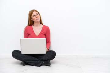 Wall Mural - Young redhead woman with a laptop sitting on the floor isolated on white background and looking up