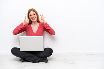 Wall Mural - Young redhead woman with a laptop sitting on the floor isolated on white background showing ok sign and thumb up gesture