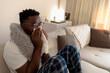 © Jelena Stanojkovic - Shot of a young man blowing his nose at home. Portrait of young black man sneezing into tissue at home. Sick african man wrapped in blanket sitting on sofa blowing his nose at home.