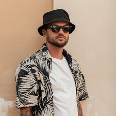 Handsome stylish hipster man with beard, sunglasses and bucket hat in fashion summer outwear with beach shirt near the wall on the street