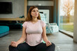 © Taras Grebinets - Serene active pregnant pretty woman sitting on lotus pose and meditating. Pregnancy and yoga, meditation concept. Caucasian beautiful woman in anticipation of a baby meditating at home.