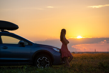 Wall Mural - Silhouette of female driver standing near her car on grassy field enjoying view of bright sunset. Young woman relaxing during road trip beside SUV vehicle