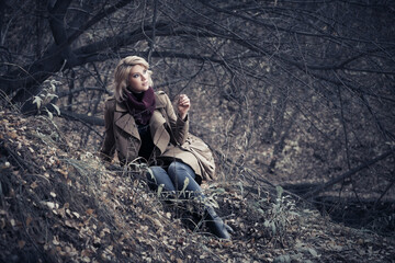 Wall Mural - Happy young fashion woman in classic beige coat sitting on ground in autumn park