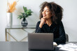 © Kateryna - Positive african american successful business woman, top manager, business consultant, dressed in a formal stylish shirt, sitting at the workplace with laptop, looking away, smiling, dreaming