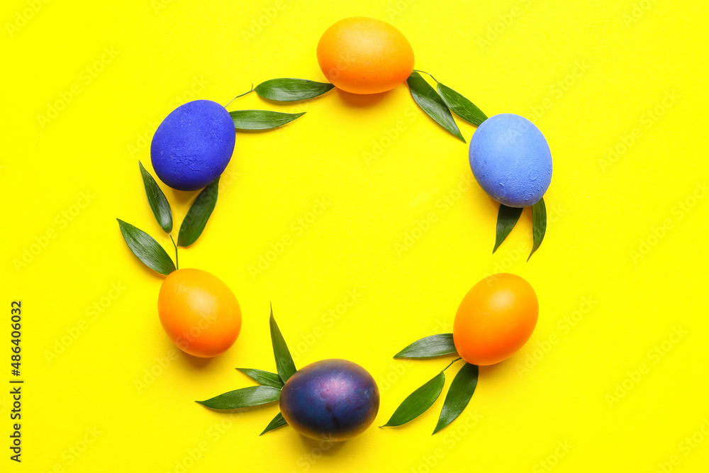 Frame made of beautiful Easter eggs and plant leaves on yellow background