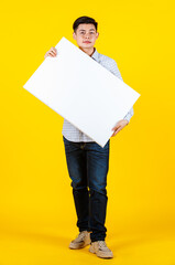 Portrait studio shot of Asian young handsome male businessman model in casual outfit standing smiling holding big white blank empty placard board for copy space advertising on yellow background