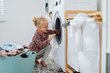 Wall Mural - Sweet pretty little girl with blonde hair, small child sits in bathroom, laundry room by large bowl full of clothes, daughter throws things into washing machine, helps mom with daily chores