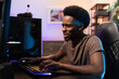 © ABCreative - Professional male pro gamer sits comfortably in chair leaning over computer, playing video games, focused on coming up with strategy, room lit with purple blue led lights