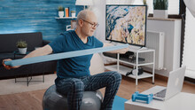 Pensioner Stretching Resistance Band To Work Arms Muscles While Watching Video Of Training Lesson On Laptop. Elder Man Using Elastic Belt And Computer, Sitting On Fitness Toning Ball.