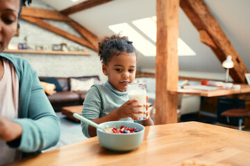 Wall Mural - Small African American girl drinks milk while having breakfast with her mother at home.