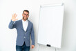 © luismolinero - Young business woman giving a presentation on white board isolated on white background saluting with hand with happy expression