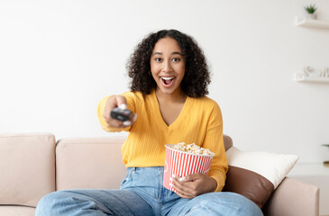 Canvas Print - Portrait of young black woman with remote control watching TV and eating popcorn on couch at home