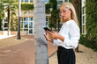 © Sergio Mena - Elegante mujer caucásica de pelo rubio con un teléfono móvil delante de un edificio empresarial