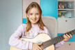 © Анна Молько - a cute little girl in a pink sweater is playing a baby guitar in her room. music training