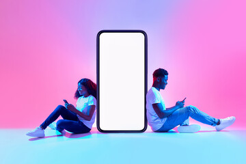 Black couple sitting near big smartphone with mockup, using gadgets, advertising new app or website in neon light