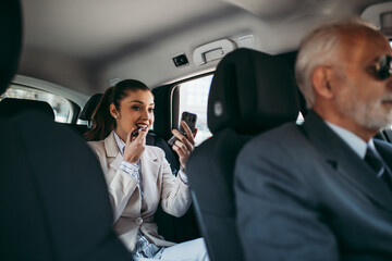 Wall Mural - Good looking young business woman sitting on backseat in luxury car and fixing her makeup. Transportation in corporate business concept.