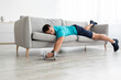Funny tired young caucasian male lies on sofa with dumbbell in living room interior, rest after training