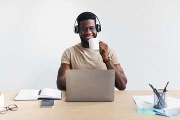 Cheerful young black guy in headphones drinking coffee, chatting online on laptop at desk against white background