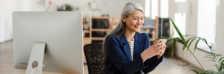 The laughing Asian woman holding a phone in her hands and looking at something in it in a light office